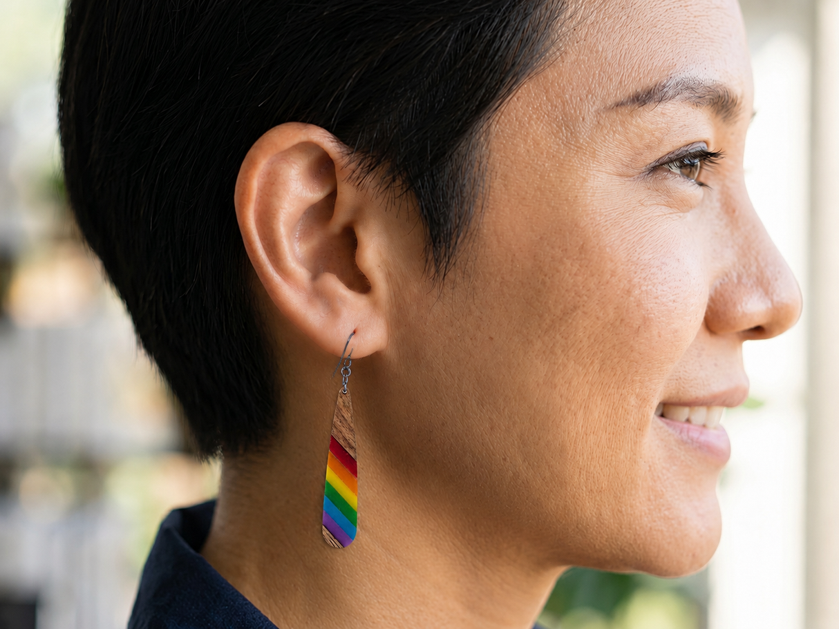Teardrop-shaped wooden earrings with diagonal stripes of pride colors and steel blue hypoallergenic ear wires worn by a nonbinary person with age in their 40s.