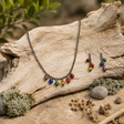 Pride necklace and earring jewelry set with glass pride-color beads, gunmetal chain, and hypoallergenic ear wires displayed on a log surrounded by greenery