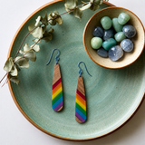 Teardrop-shaped wooden earrings with diagonal stripes of pride colors and steel blue hypoallergenic ear on a round sage colored plate with eucalyptus a a small bowl of gemstones in blues and greens.