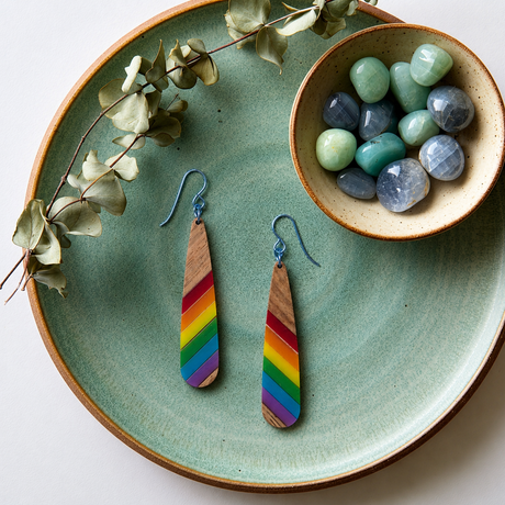 Teardrop-shaped wooden earrings with diagonal stripes of pride colors and steel blue hypoallergenic ear on a round sage colored plate with eucalyptus a a small bowl of gemstones in blues and greens.