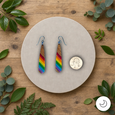 Teardrop-shaped wooden earrings with diagonal stripes of pride colors and steel blue hypoallergenic ear wires on a round beige stone with a quarter for scale, surrounded by green leaves on a wooden surface.