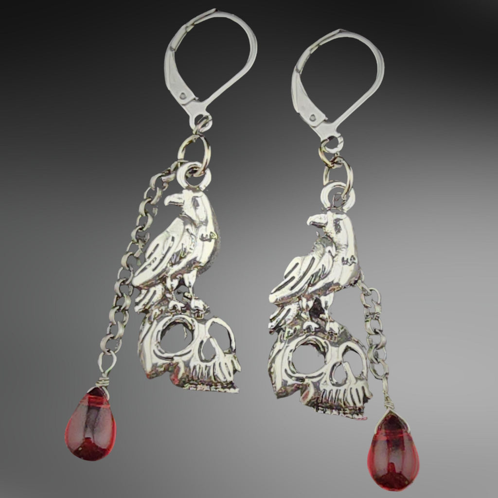 Silver-toned earrings, each with a  a raven perched menacingly atop a human skull and a blood-red glass bead dangling below on a silver-toned chain.