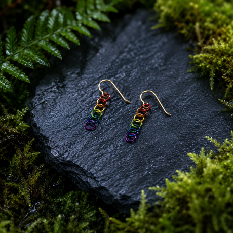 Rainbow chain link earrings with gold-filled ear wires on a dark stone surface surrounded by green foliage