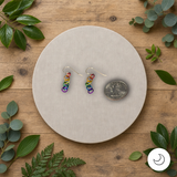 Gold-filled pride earrings with rainbow rings on a round beige mat with a coin for scale, surrounded by green leaves on a wooden surface.