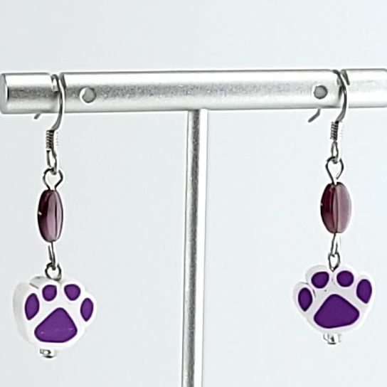 Earrings, purple paw prints on white paw print background. Rice-shaped purple bead above each print. Ear wires are silver tone.